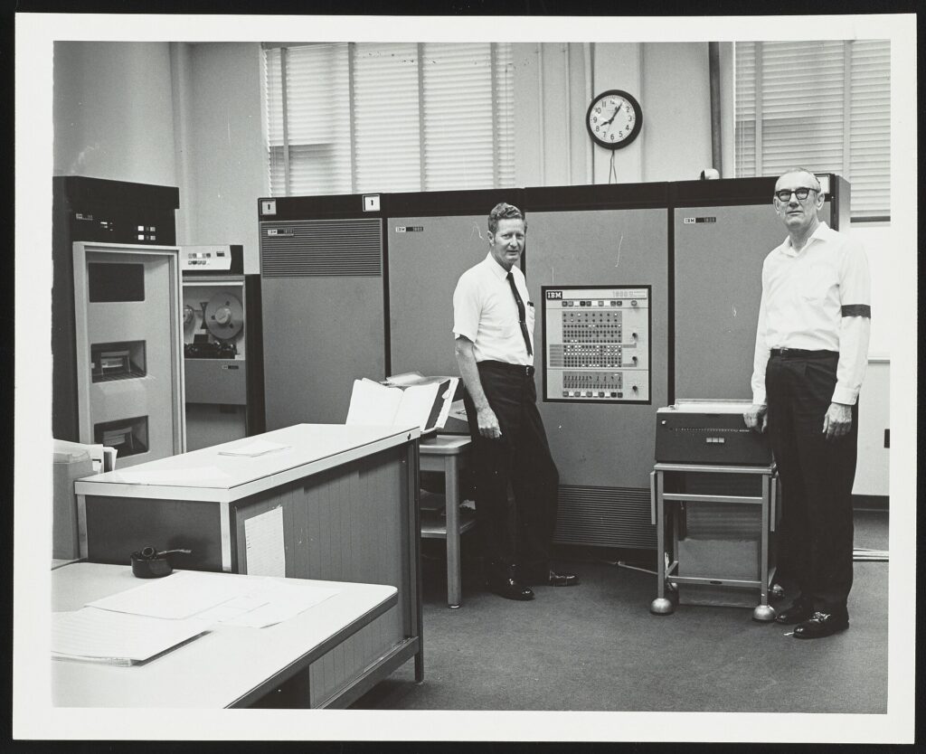 View of the IBM 1800 Data Acquisition and Control System computer used to accept and process data from a mass spectrometer at the Exxon Research and Engineering Company laboratory in Baytown, Texas. This photograph was taken on the day that use of the computer was discontinued at the laboratory. Henry Earle Lumpkin (Research Associate in charge of computer operations) stands to the left while laboratory technician Jack L. Taylor is visible on the right. Taylor is wearing a black band on his right arm to mark the "death" of the computer.