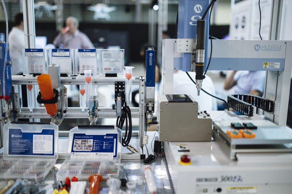A robotic arm is positioned next to various tools and supplies used in laboratory settings. Attendees at the technology event observe the equipment demonstrating its functions.
